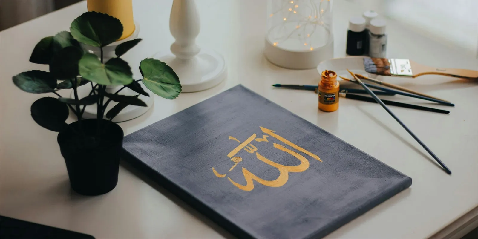 Overhead shot of a white glossy table with a painted canvas on which 'Allah' is written in gold color, There's a lit yellow candles, a plant, a showpiece, and painting tools near it.